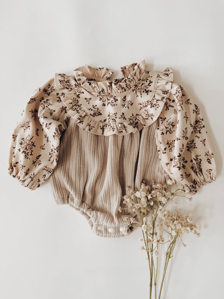 Infant romper with neutral color floral print on top with a ruffled collar. The bodice is a taupe ribbed fabric with buttons at the crotch.