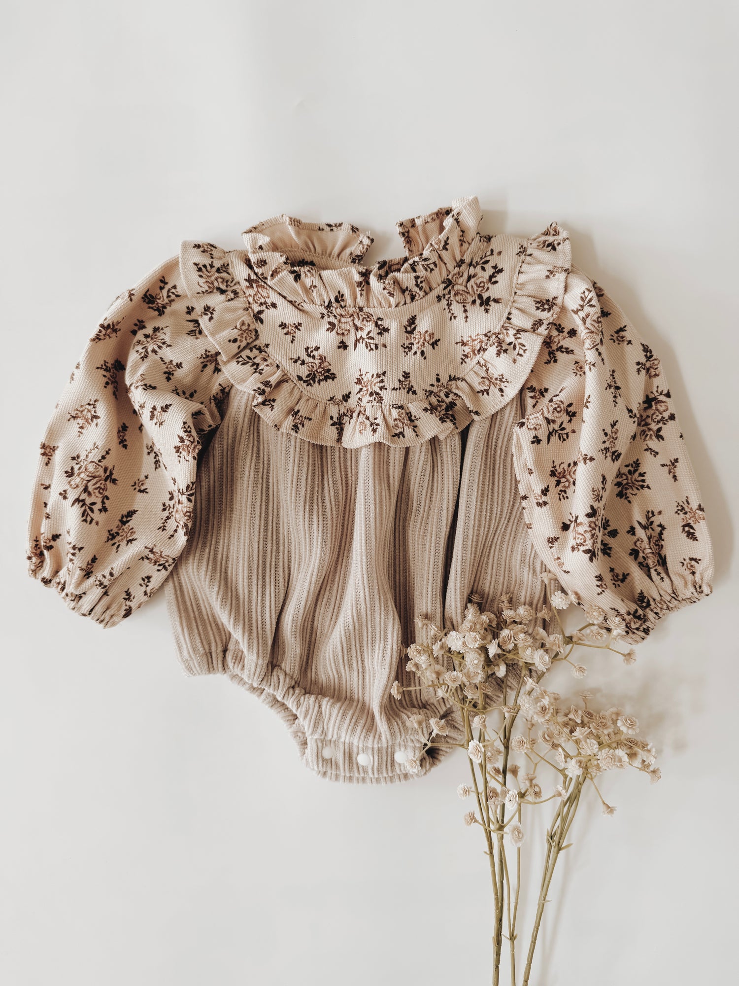 Infant romper with neutral color floral print on top with a ruffled collar. The bodice is a taupe ribbed fabric with buttons at the crotch.  