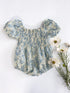 Ivory cotton baby romper with blue toile floral print, featuring puff sleeves, a smocked bodice, and elastic leg openings, styled flat lay with white flowers.