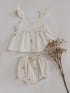 Ivory two-piece baby outfit made from duck cotton fabric featuring delicate blush pink floral embroidery, flutter strap top with buttons, matching bloomers, pink socks, and beige baby shoes laid flat on a neutral background.