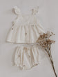 Ivory two-piece baby outfit made from duck cotton fabric featuring delicate blush pink floral embroidery, flutter strap top with buttons, matching bloomers, pink socks, and beige baby shoes laid flat on a neutral background.