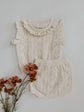 Cream-colored knitted baby outfit with ruffled collar on a light gray background