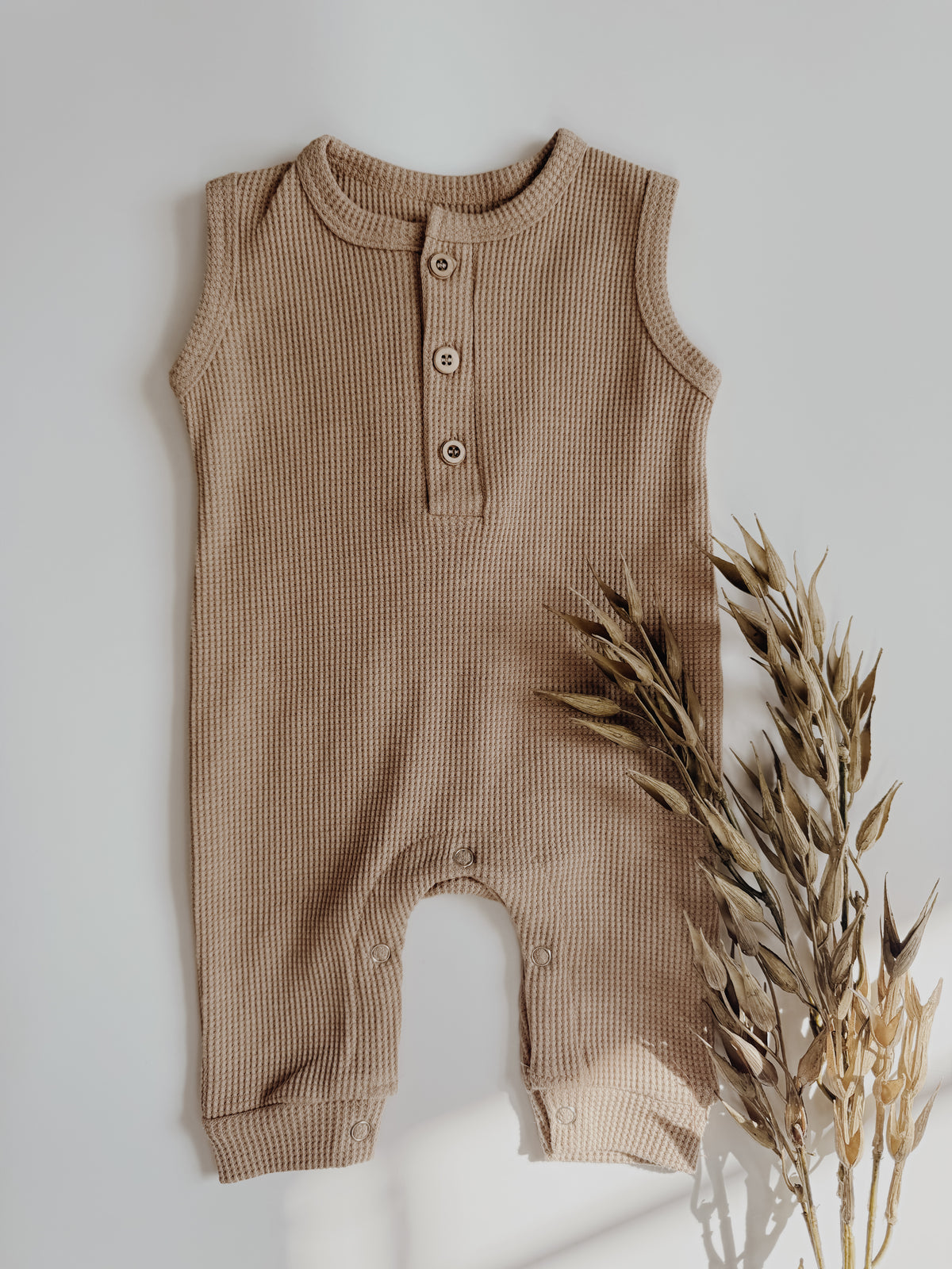 A sleeveless ribbed knit baby romper in a warm taupe-brown color, styled flat-lay on a light gray background with dried grass stems.