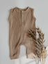 A sleeveless ribbed knit baby romper in a warm taupe-brown color, styled flat-lay on a light gray background with dried grass stems.